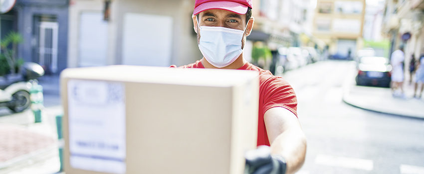 Young delivery man wearing uniform and coronavirus protection medical mask holding cardboard package at town street.